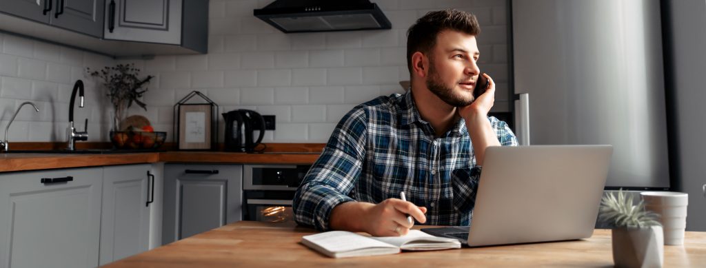 Happy remote worker on a phone call and using his laptop at the kitchen table